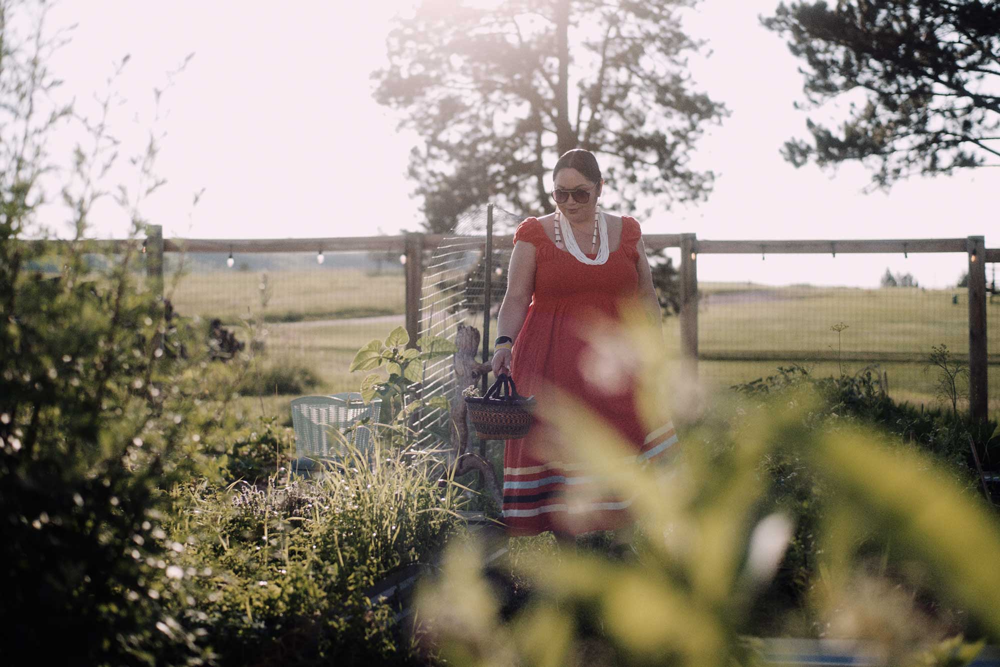 A woman, Kimberly Tilsen-Brave Heart, wearing a red sundress walks through a garden holding a basket.