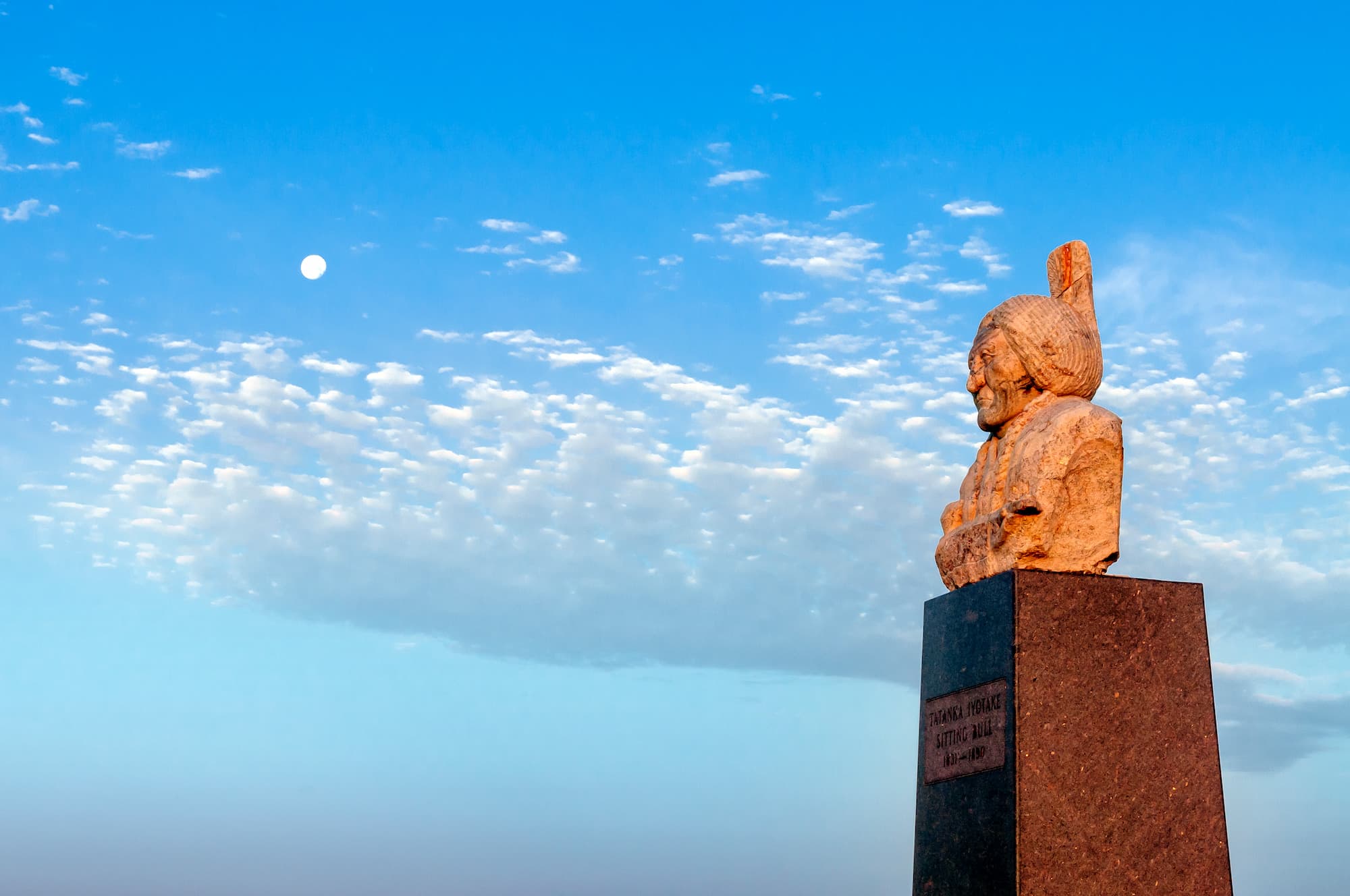 A sculpted bust of Sitting Bull looks out the horizon in front of a blue sky with white clouds