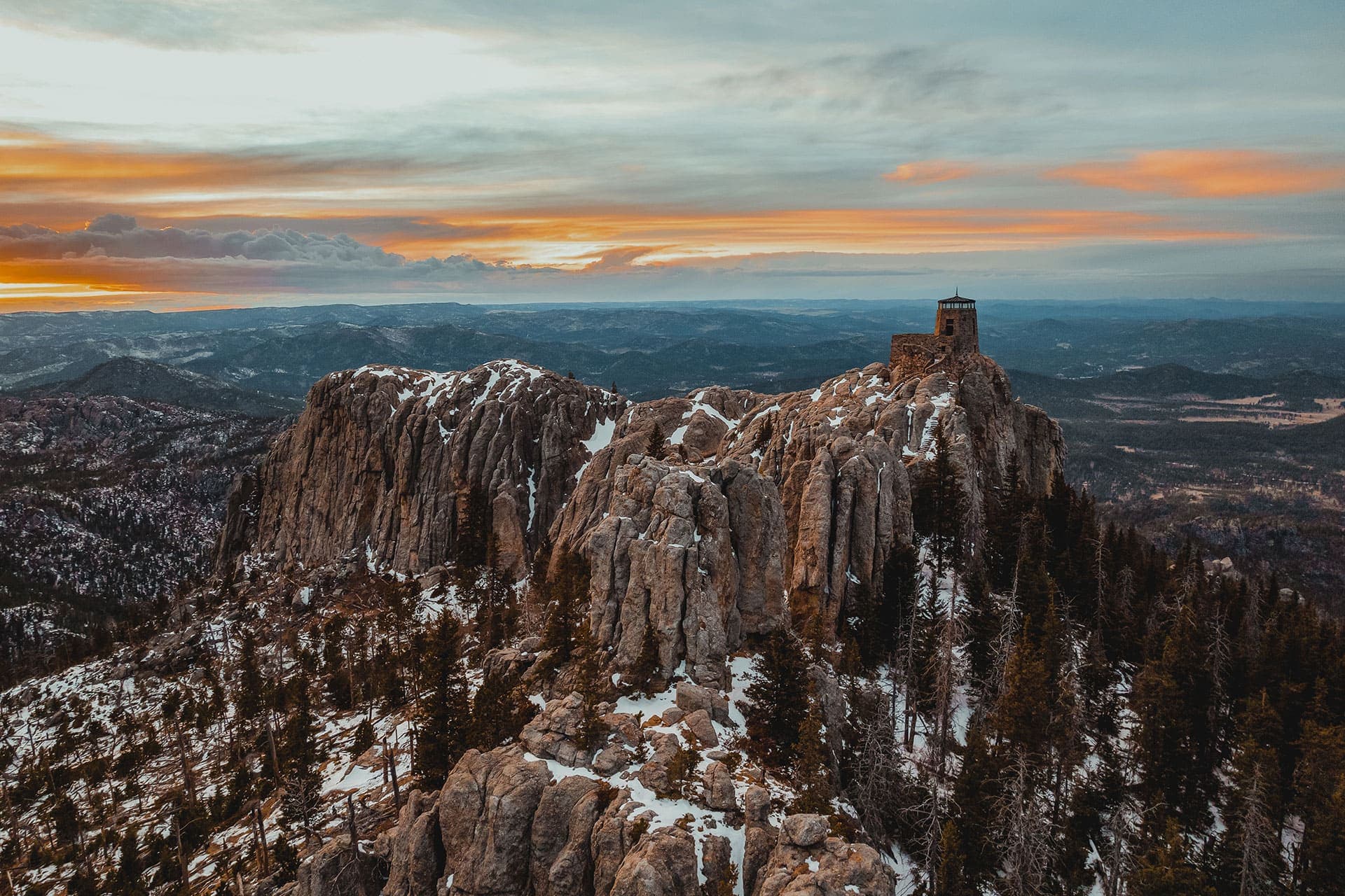 Aerial view of Black Elk Peak in South Dakota, jagged rock formations with a dusting of snow and a sunset in the background.