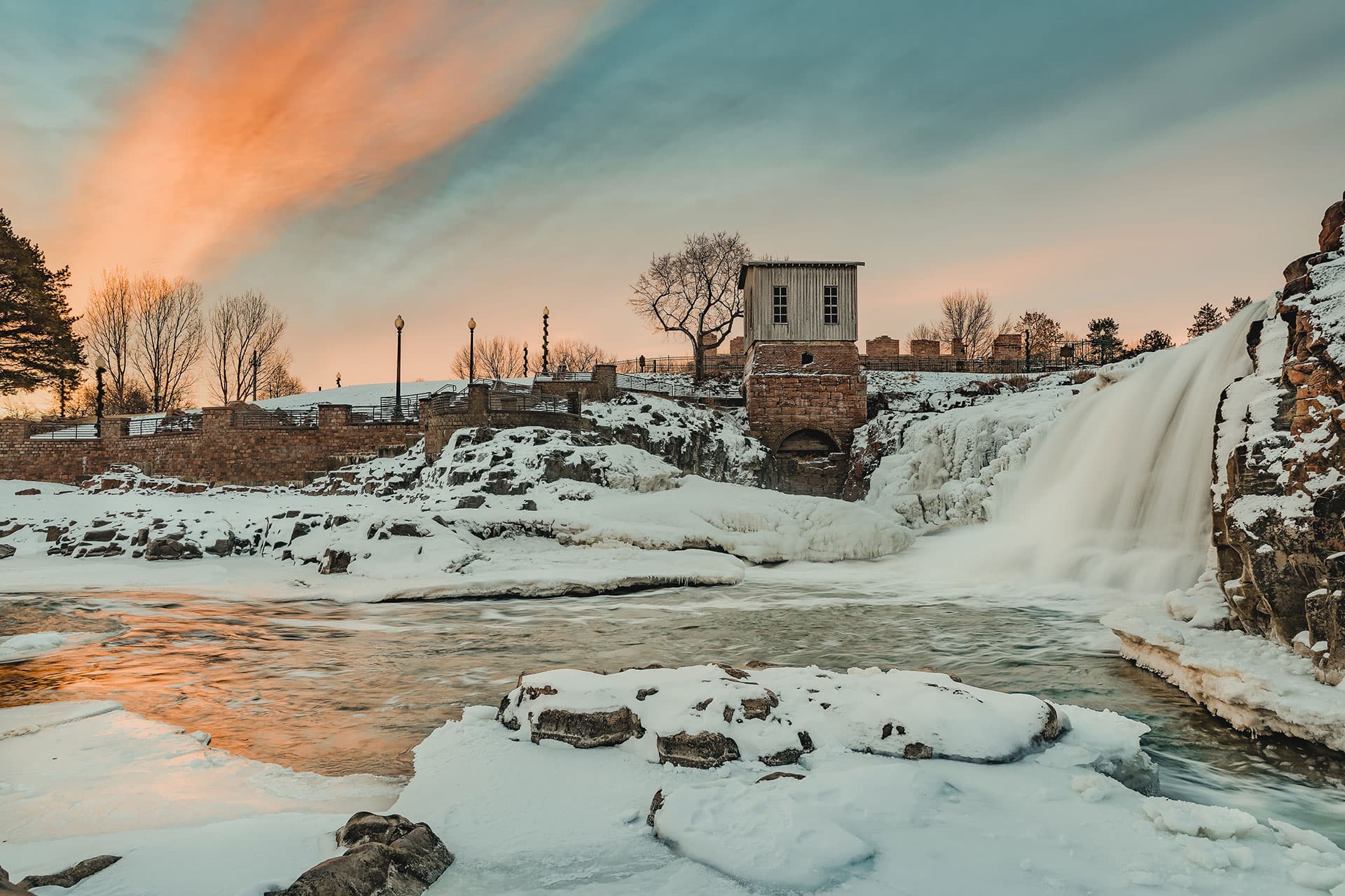 A frozen river landscape with snow and a waterfall with a stone bridge and pink sunset in the background at Falls Park in Sioux Falls, South Dakota.