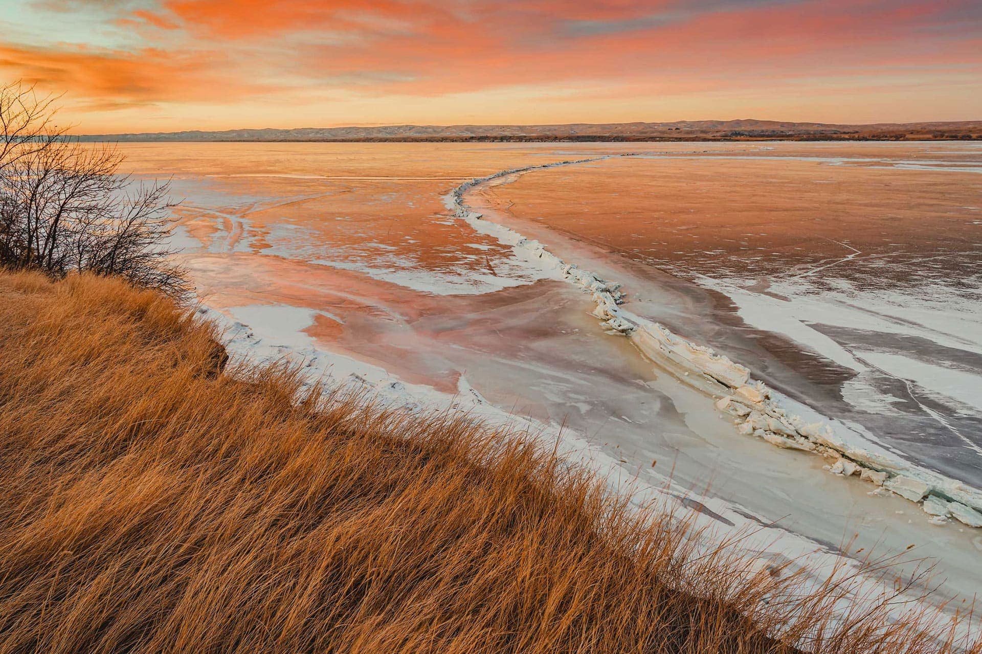 Scene of the Missouri River in South Dakota frozen with ice during a pink and orange sunset with tall grass along the riverbank.