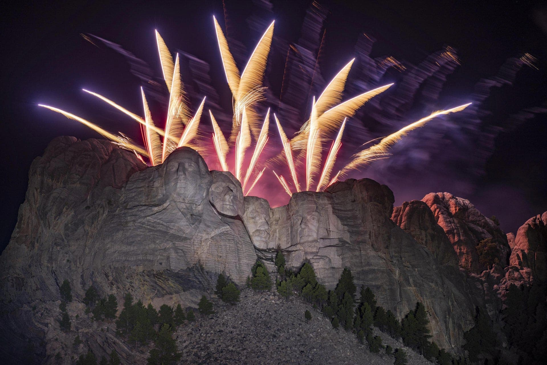 Fireworks explode over Mount Rushmore National Memorial, with the face of the sculpture illuminated by their multicolored light.