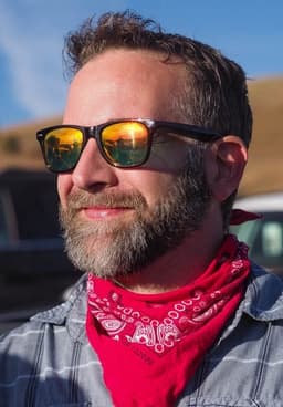 A man smiles as he looks into the sun while wearing sunglasses and a red handkerchief around his neck