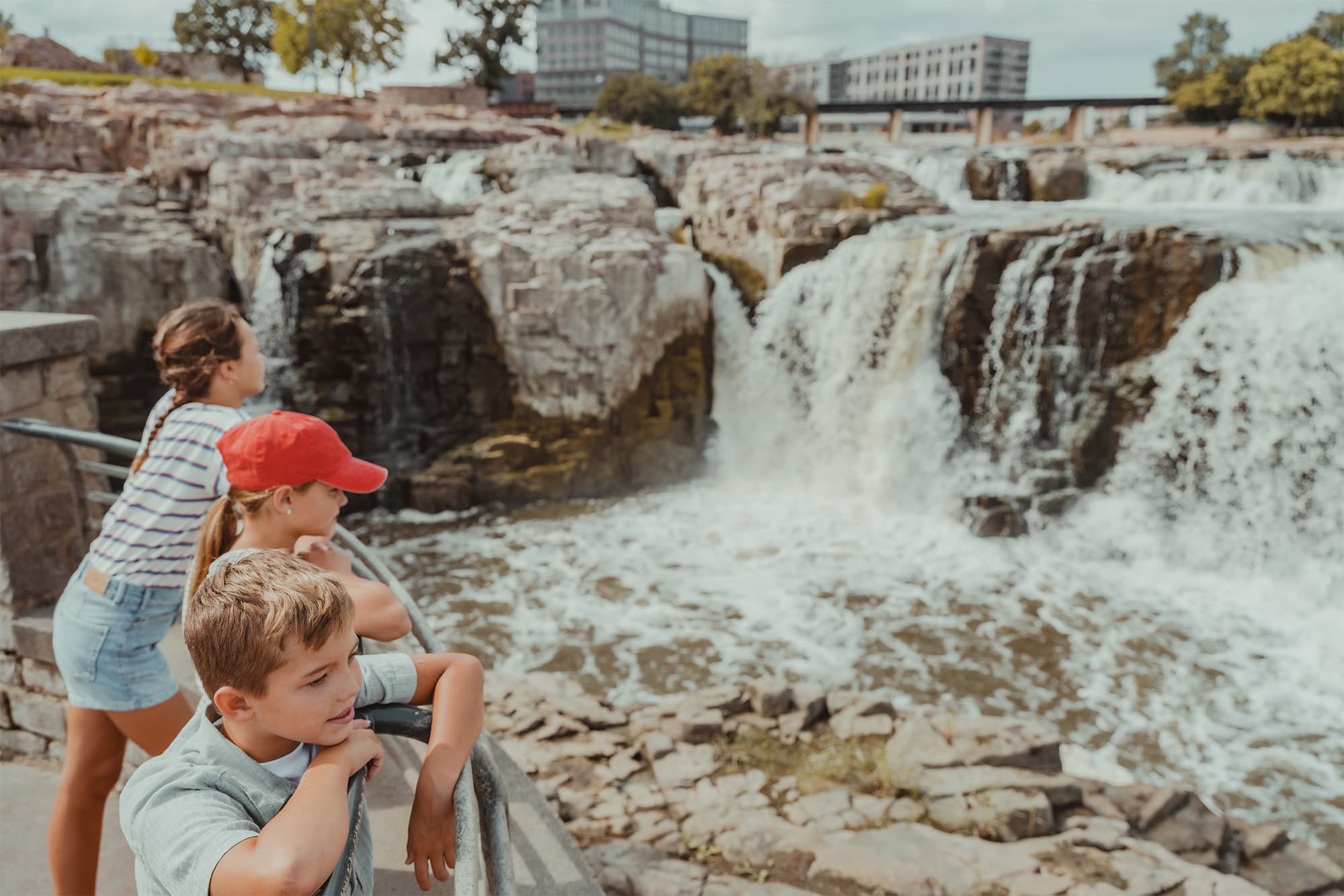 Three children stand on a lookout above Falls Park, watching the rushing water on a sunny day.