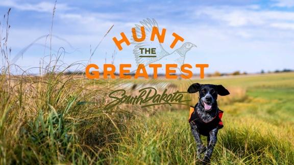 A black dog runs toward the camera in a field beside the words HUNT THE GREATEST SOUTH DAKOTA