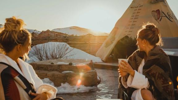 to women sitting outside a teepee in winter in spa robes with hot drinks