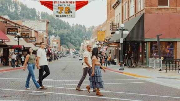 Two couples cross a city street in Deadwood, South Dakota. A banner for the city's Days of '76 celebration hangs overhead.