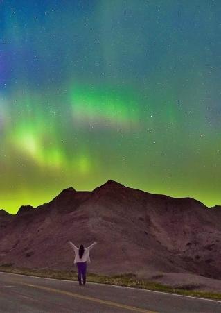 A woman raises her hands in the Badlands under the Northern Lights