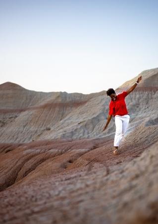 A woman in red shirt and white pants holds her arms out with joy as she walks through Badlands National Park