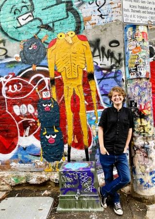 Artist Logan Beert stands in a black shirt and blue jeans in front of his alien-like creatures painted in Art Alley