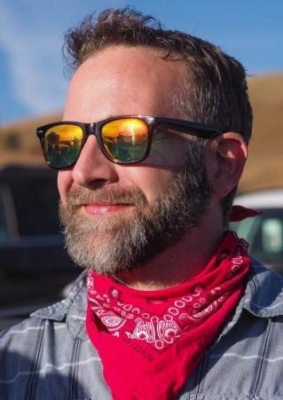 A man smiles as he looks into the sun while wearing sunglasses and a red handkerchief around his neck