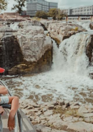 Three children stand on a lookout above Falls Park, watching the rushing water on a sunny day.
