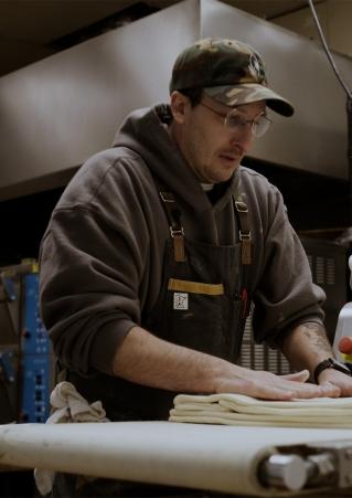 Peter Mitchell, wearing wire-rimmed glasses and a camo-print baseball cap, rolls layers of croissant dough in the bakery.