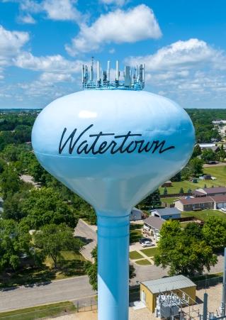 An aerial view of the Watertown water tower in Watertown, South Dakota, against a bright blue sky with fluffy white clouds.