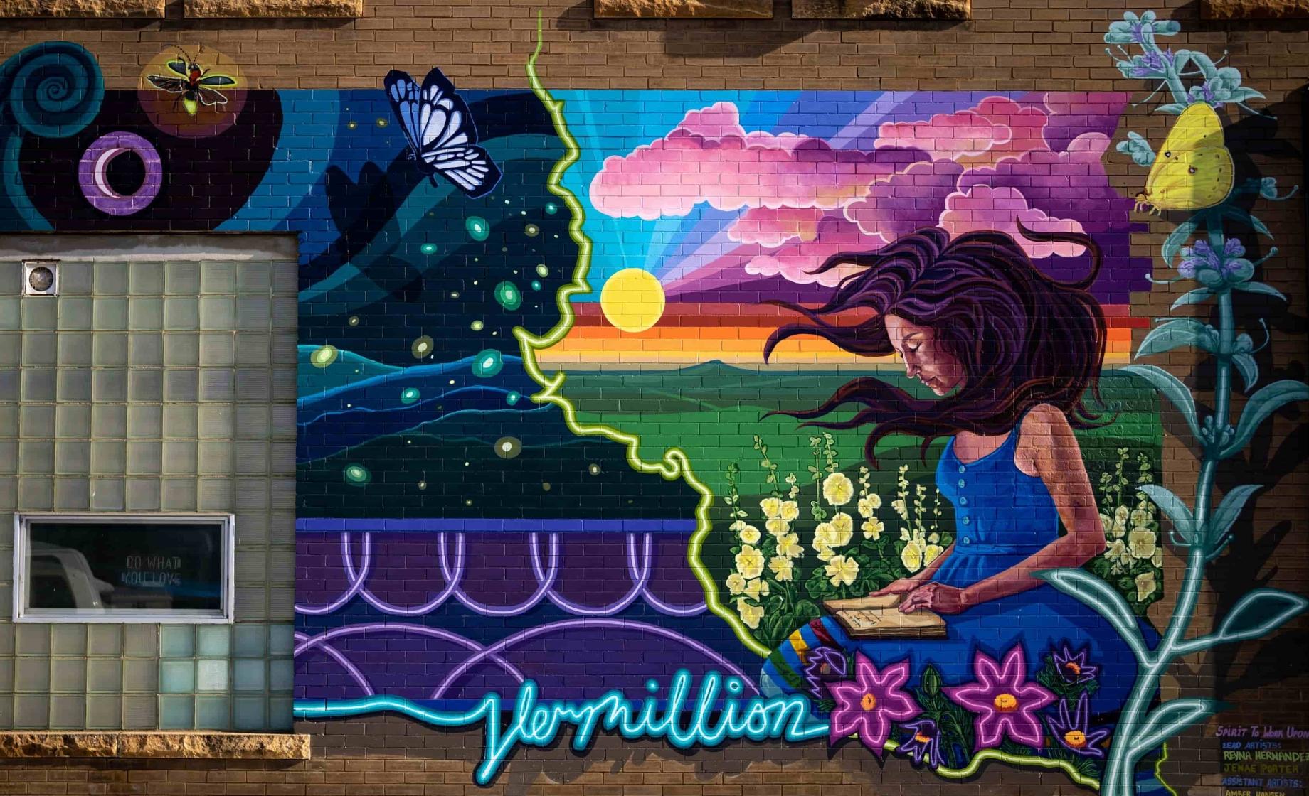 A mural in the shape of South Dakota features a woman looking at flowers while a sun shines through purple clouds -- this is painted on the outside of a brick building in Vermillion