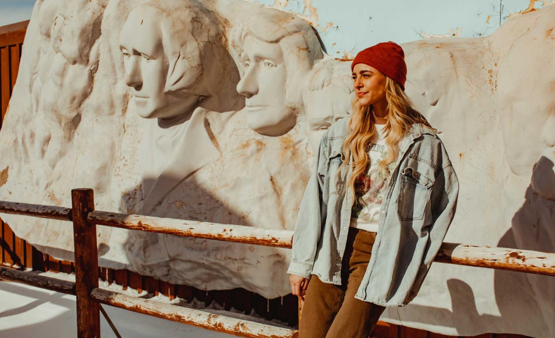 Woman in a denim jacket and read beanie leaning against a fence in front of a cartoonish replica of Mount Rushmore.