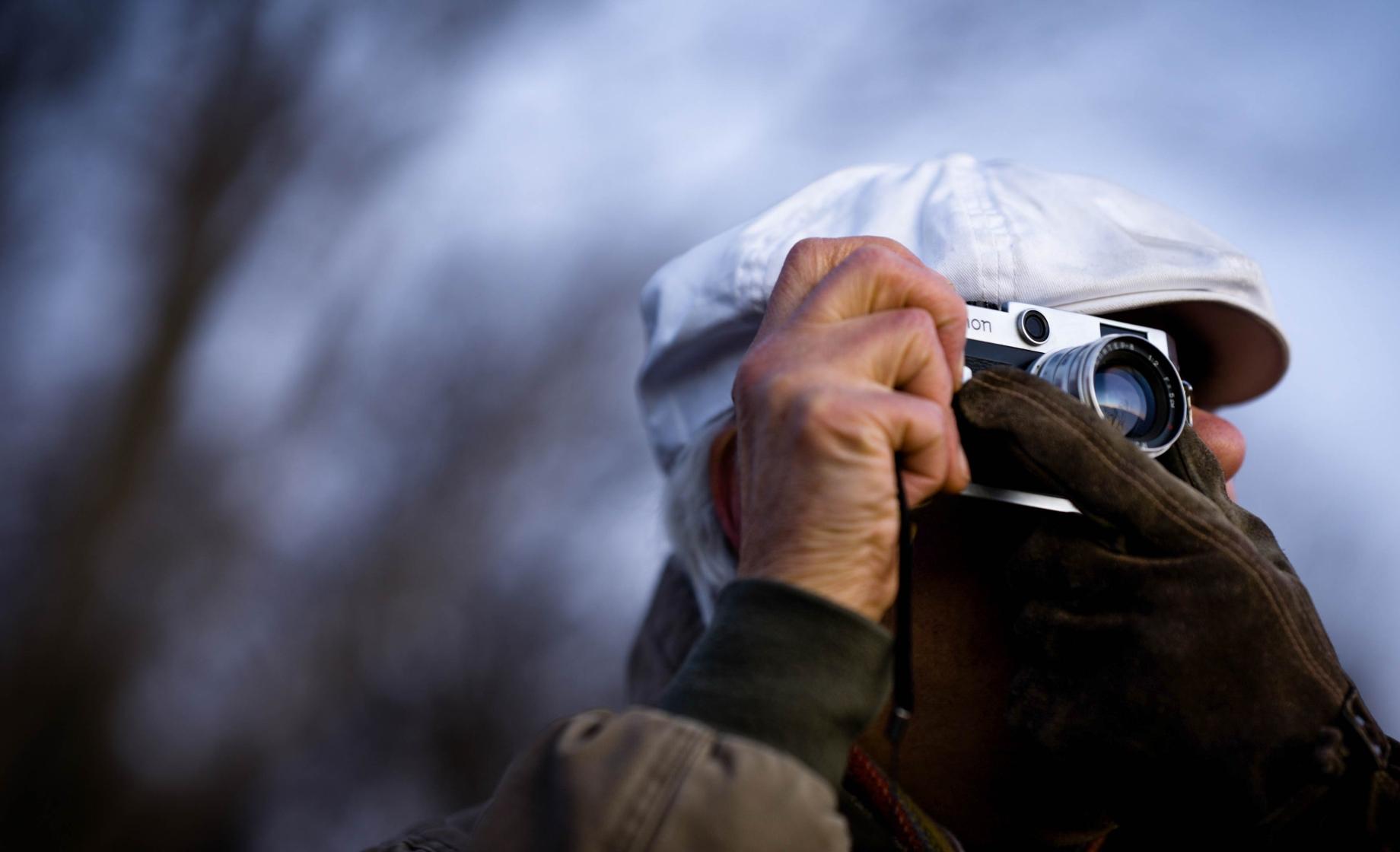 Close up image off a man holding a camera to shoot, with bare trees blurred in the background