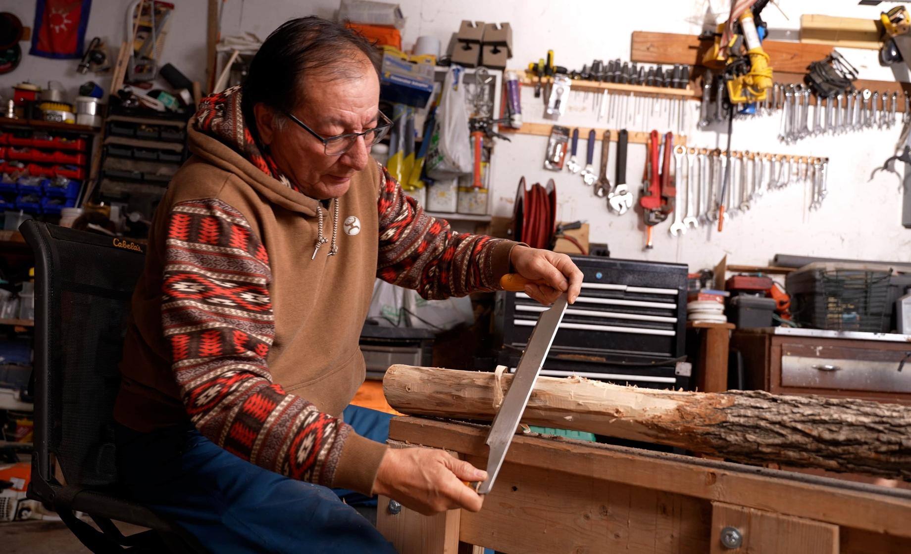 An Indigenous man carves a piece of wood that will become a traditional bow