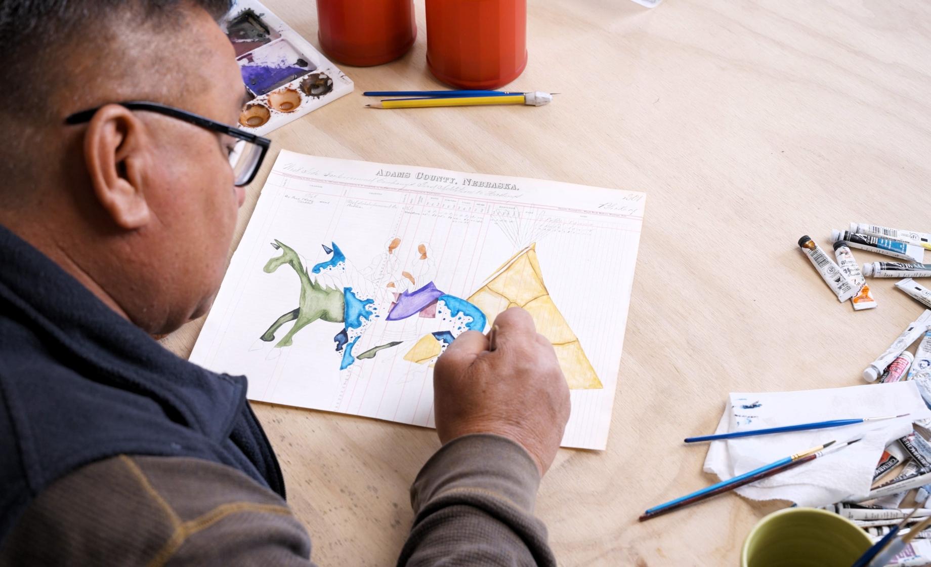 An Indigenous man makes colorful art on ledger paper