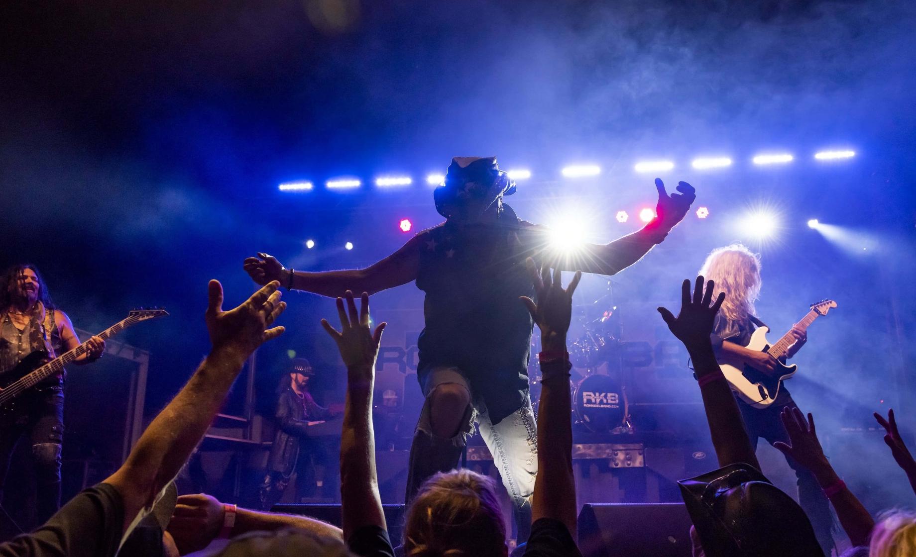 A singer poses onstage with a guitarist under concert lights in front of a happy crowd