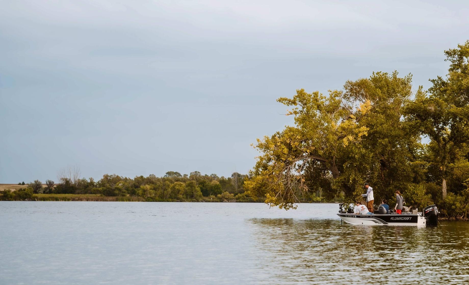 Anglers sit and stand in a boat in front of a tree while fishing open water in NE South Dakota