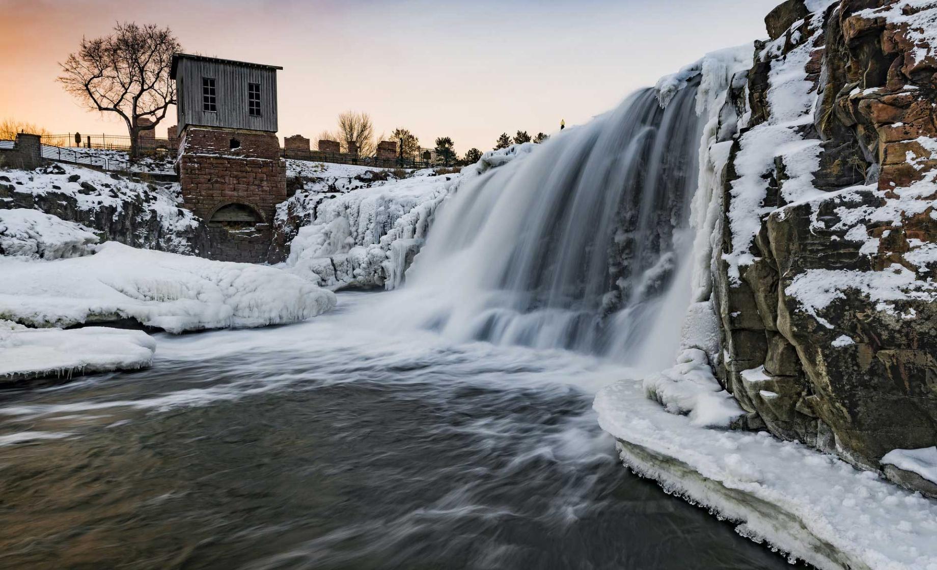 A waterfall at Falls Park in Sioux Falls, Sioux Dakota, is pictured at sunset surrounded by snowy banks.