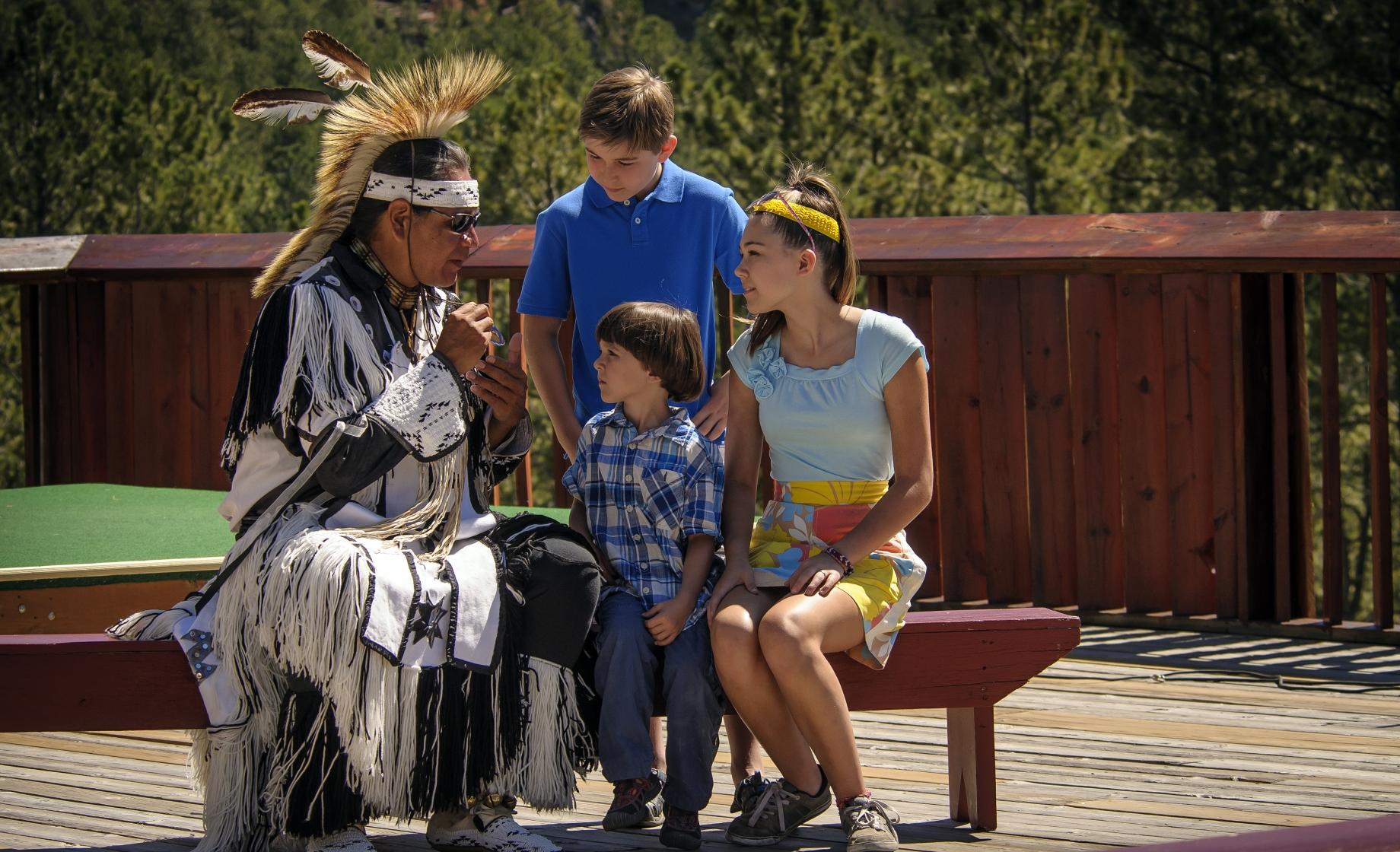 A Native American storyteller and performer in powwow regalia talking with a group of kids at Crazy Horse Memorial