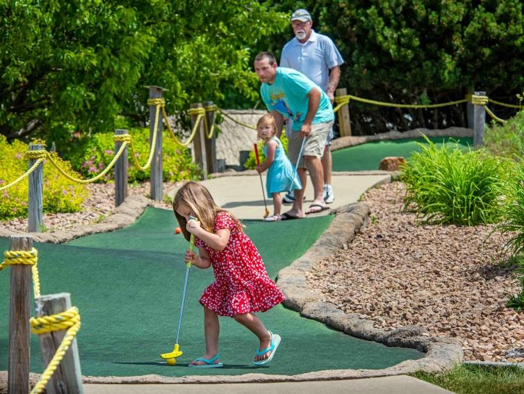 A family looks on as a young child awkwardly knocks her mini golf ball into the hole