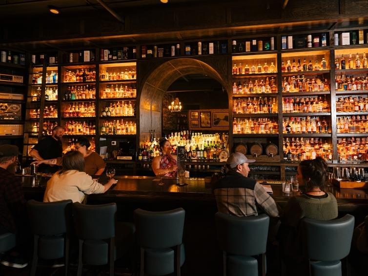The Windsor Block Bar in Rapid City, South Dakota, with couples chatting on barstools and a half-wall of shelves filled with liquor bottles lit from behind.