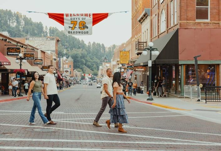 Two couples cross a city street in Deadwood, South Dakota. A banner for the city's Days of '76 celebration hangs overhead.