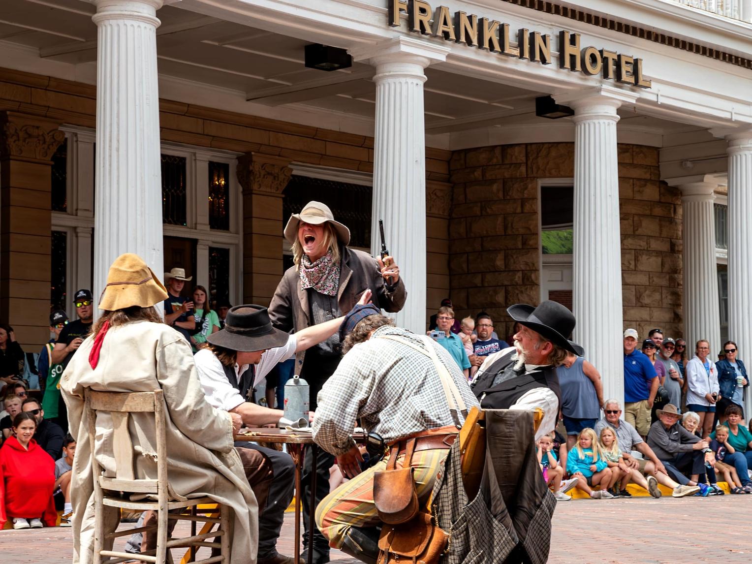 An actor portraying an Old West gunman confronts a group of actors at a table on Deadwood's Main Street while people look on
