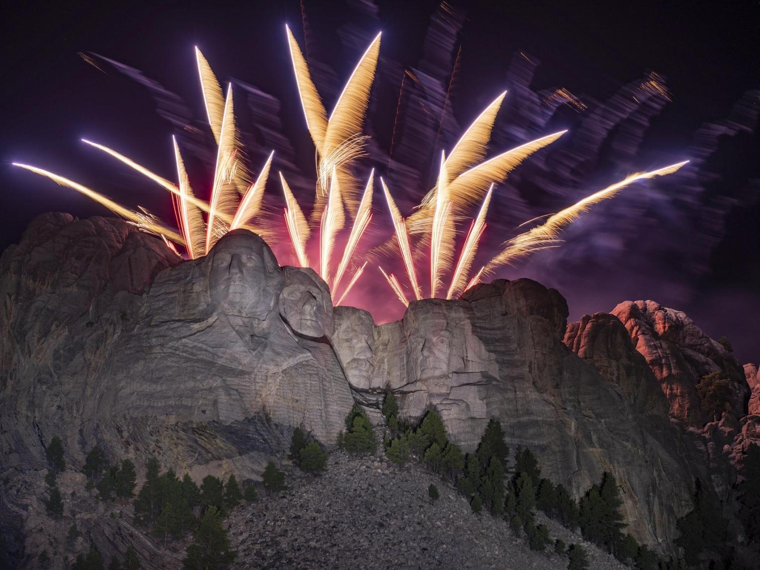 Fireworks explode over Mount Rushmore National Memorial, with the face of the sculpture illuminated by their multicolored light.