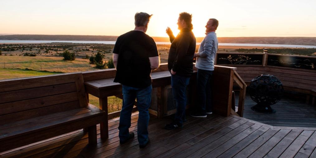 Three men smile and laugh together on a deck overlooking a vast grassland in South Dakota at sunset.