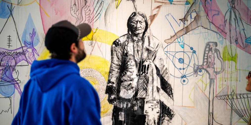 A person wearing a blue sweatshirt and black baseball cap views art of an Indigenous leader.