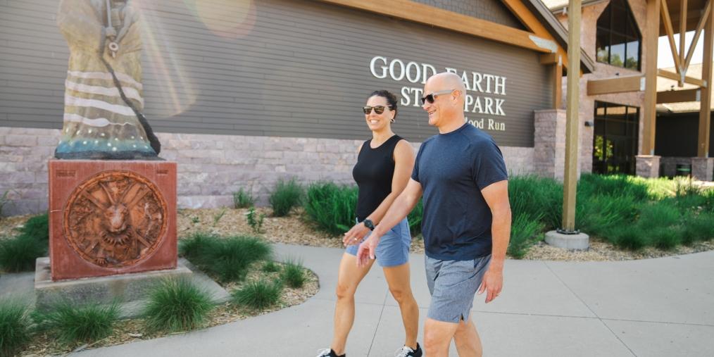 Two adults wearing shorts, sunglasses and dark-colored shirts walk in front of the Good Earth State Park visitor center.