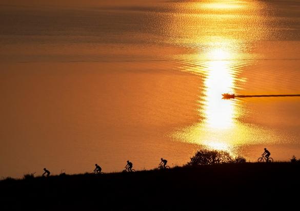 Five mountain bikers ride the Oahe West Shore Bike Trail as the sun sets on Lake Oahe in the background, with a fishing boat heading to dock racing alongside.