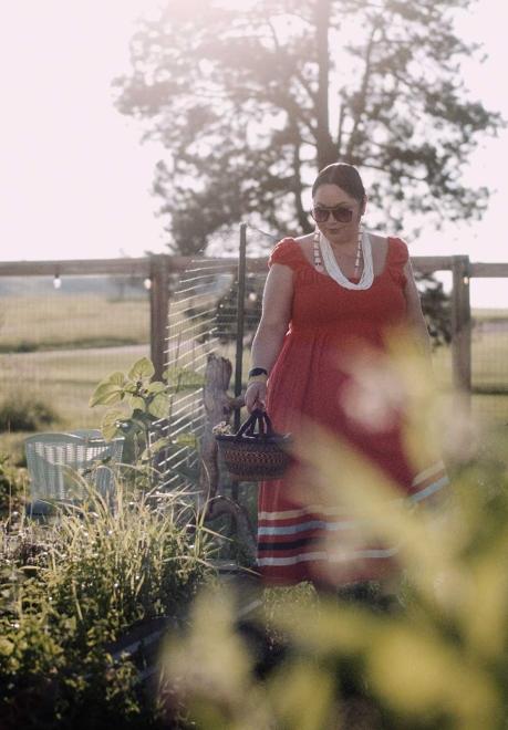 A woman, Kimberly Tilsen-Brave Heart, wearing a red sundress walks through a garden holding a basket.