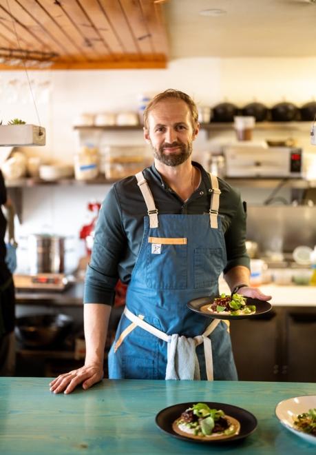 Portrait of Skogen Kitchen in Custer's owner and chef, Joseph Raney with kitchen in the background and finished plates on a vintage green countertop 