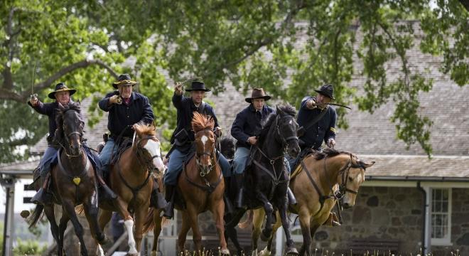 Cavalry reenactors on horseback ride toward the camera with historic buildings in the background at the Fort Sisseton Historical Festival. Fort Sisseton Historic State Park, South Dakota.